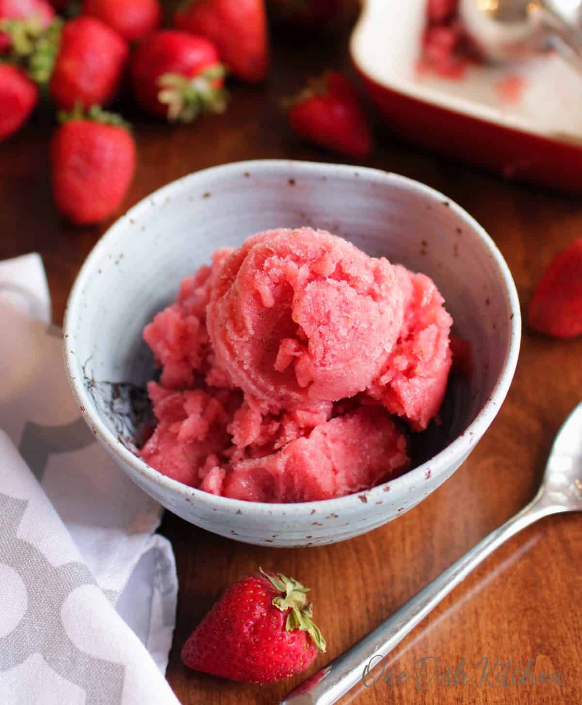 an overhead view of a bowl of strawberry sorbet next to strawberries scattered around a table.
