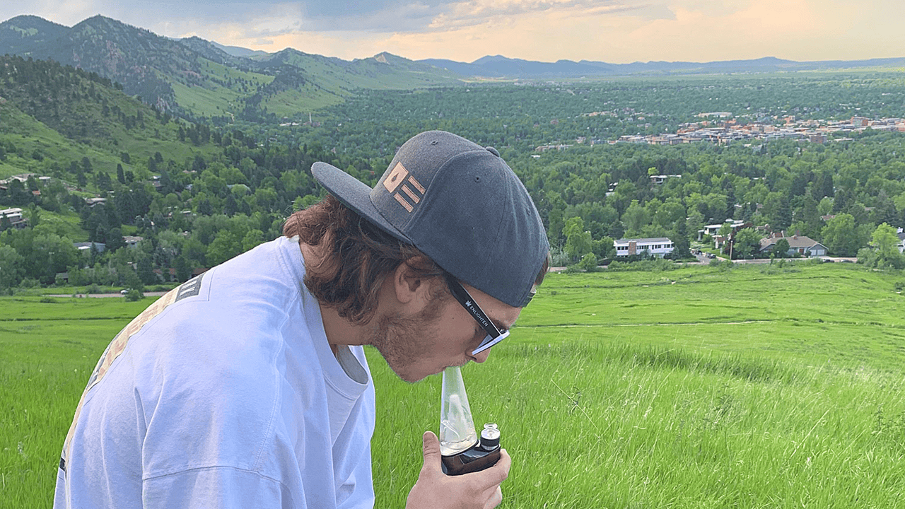 man wearing hat takes CBD dab with mountainscape in background