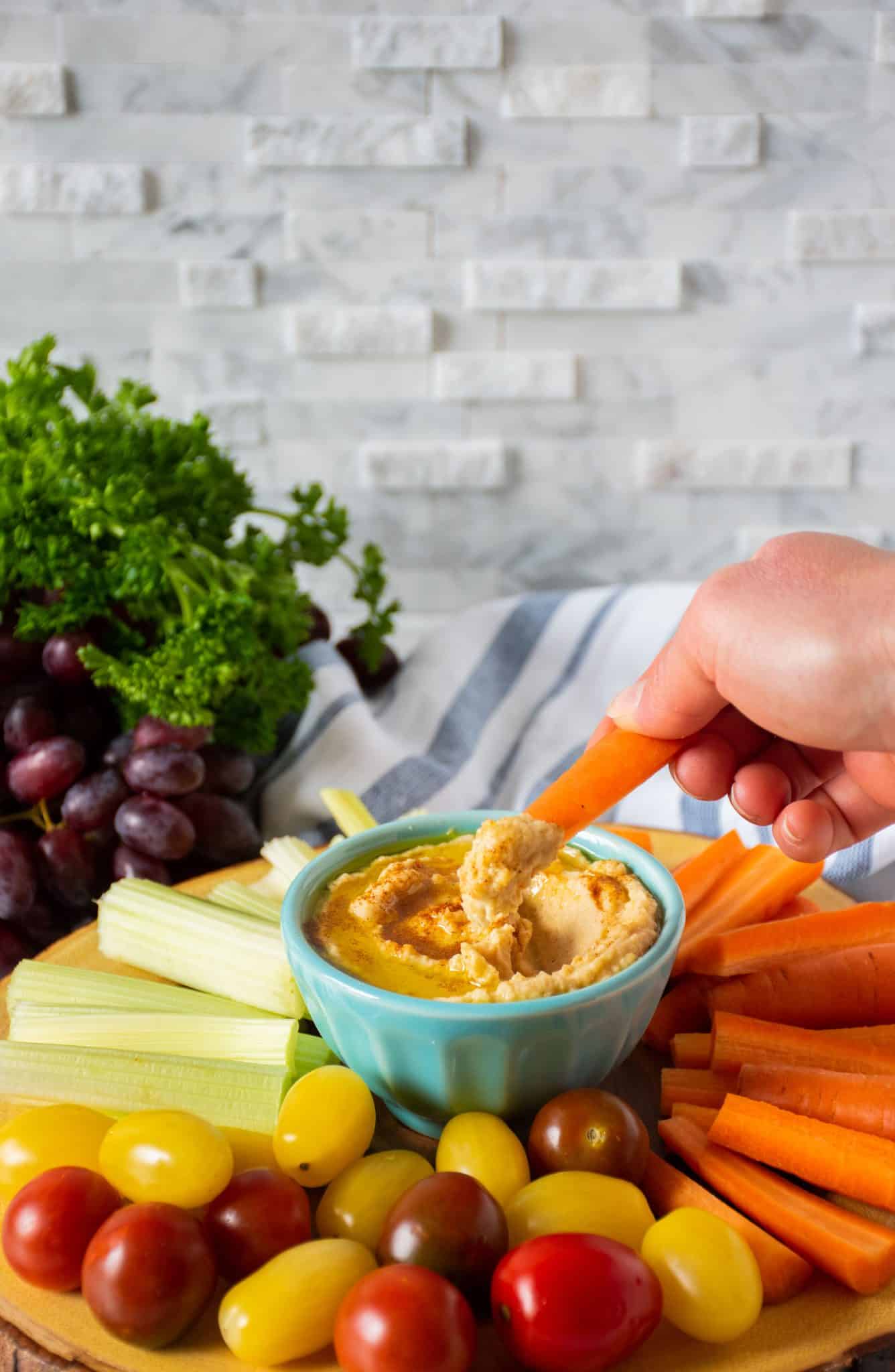 Bowl of pantry basics hummus in the middle of a vegetable platter, with a hand dipping a carrot stick in the hummus.