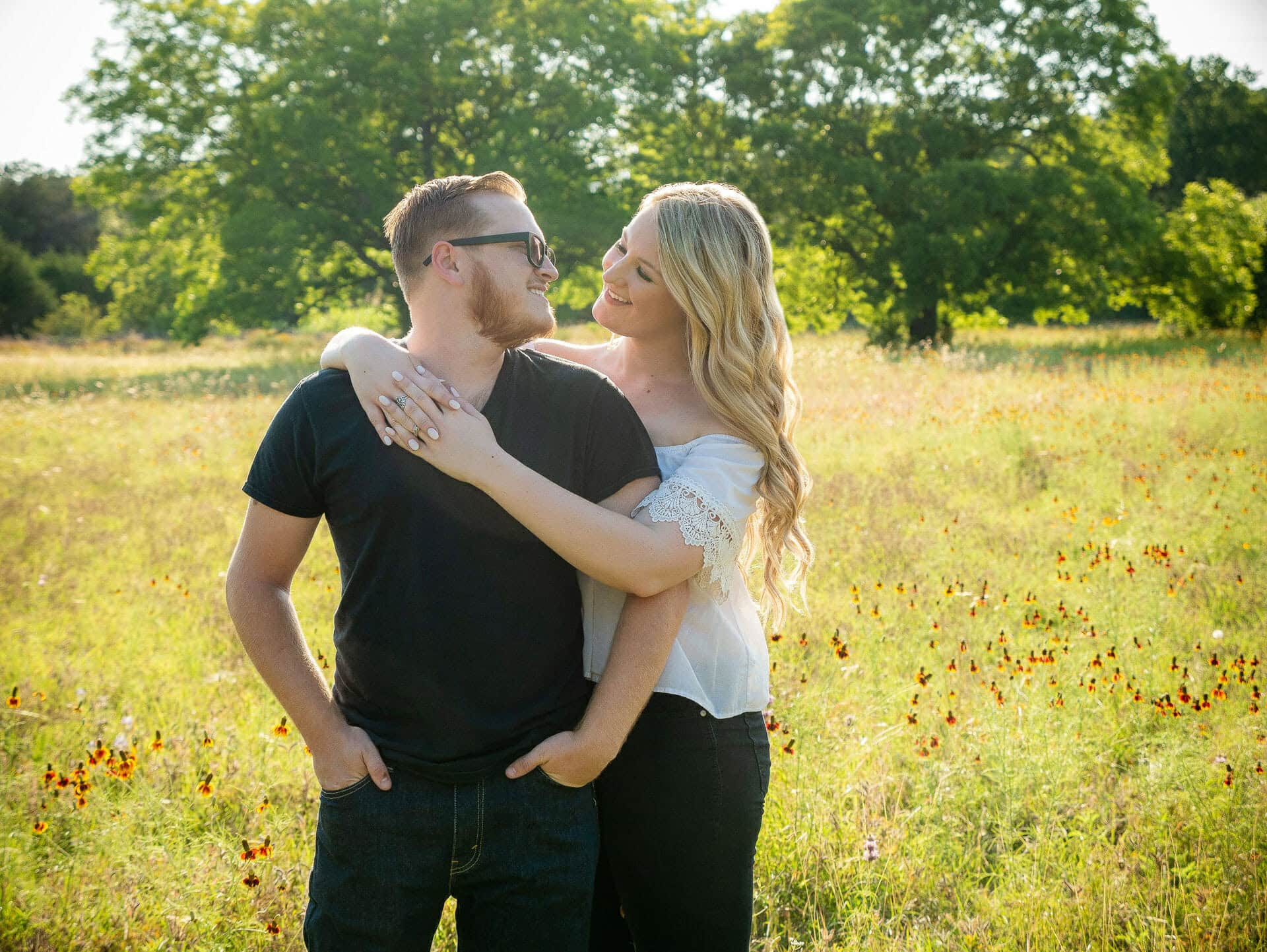 engagement photos of a man in a black shirt wearing glasses and a female blonde in a white shirt and jeans standing in a field during sunset