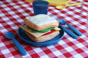 Photo of a pretend sandwich on a blue plate. The plate is on a red checkered table cloth in a dramatic play center kitchen.