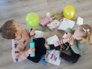 Ein Junge sitzt auf dem Boden und bläst einen Luftballon mit einer Luftpumpe auf. Ein Mädchen sitzt neben dem Jungen auf dem Boden und bläst in eine Rolle aus Geschenkpapier. Neben den Kindern liegen Geschenke, ein Luftballon, Girlanden und Konfetti.