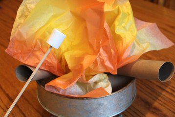 Photo of a pretend campfire with orange and yellow tissue paper for flames. Paper towel tubes are used as logs. A campsite is a great summer dramatic play idea.