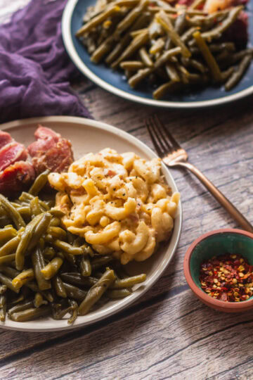 a close up of southern style green beans on a white plate with macaroni and cheese and some red pepper flakes