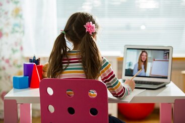 Photo of you girl sitting at a desk at home - engaged in a distance learning reading lesson. She faces a computer that shows a teacher standing in front of an easel.