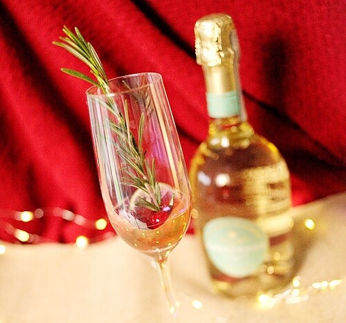 A champagne glass filled with raspberry liqueur, rosemary sprig, fresh cranberries and a slice of lemon for the Raspberry Rosemary Fizz
