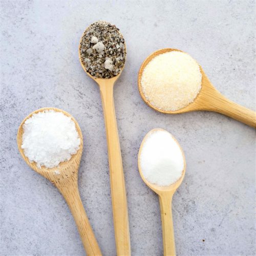 4 wooden spoons on a pale background each with a different type of salt. Sea salt, flavoured, table & garlic.