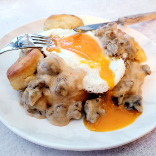 Wide shot of gravy and biscuits with a runny fried egg. Cutlery placed as being eaten.