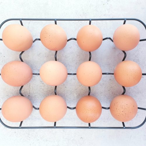Overhead shot of 12 pale eggs in their shell on a pale blue background sitting in a dark grey wire rack.