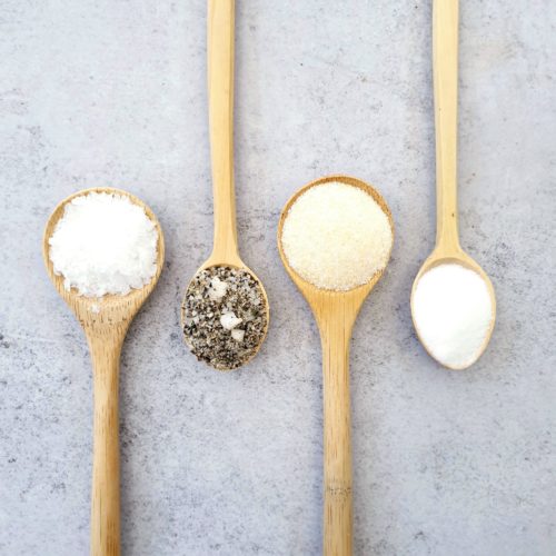 4 wooden spoons on a pale background each with a different type of salt. Sea salt, flavoured, table & garlic. Spoon are lined up with 2 pointing to the top and 2 to the bottom.
