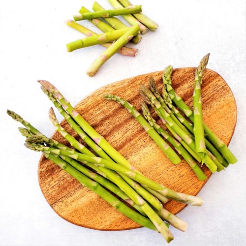 Asparagus show on a wooden board. Some are whole and others are prepared by snapping. The discarded sections are show to the side.