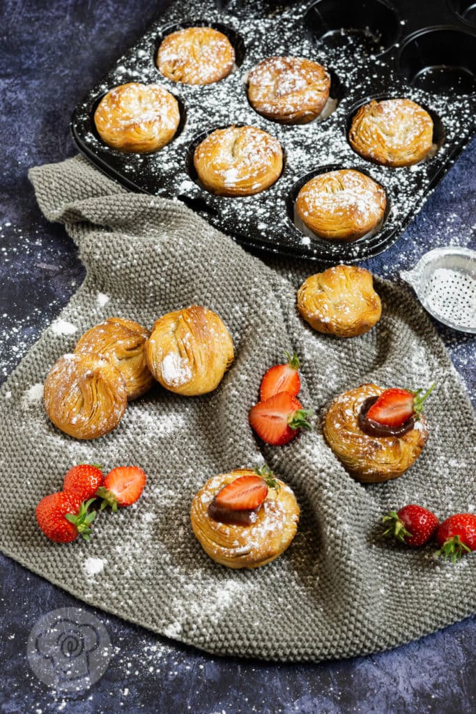 Cruffins - Croissant trifft Muffin 2 Cruffins von oben fotorgafiert, teilweise im Muffinblech, teilweise auf einem grauen Tuch