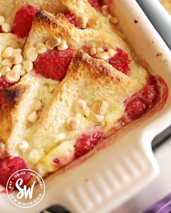 Raspberry Bread and Butter Pudding studded with white chocolate chips in a Le Creuset stoneware dish.