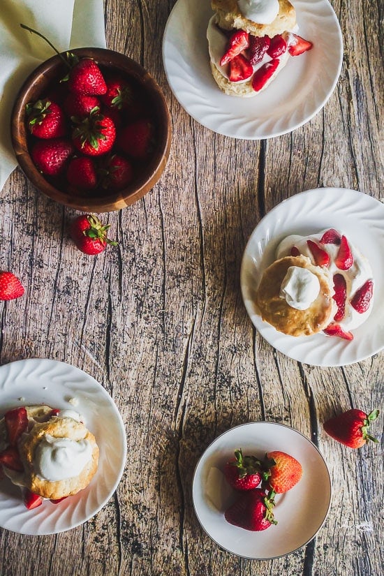 overhead shot of homemade strawberry shortcakes on plates with a bowl of strawberries and strawberries strewn about on a wooden table