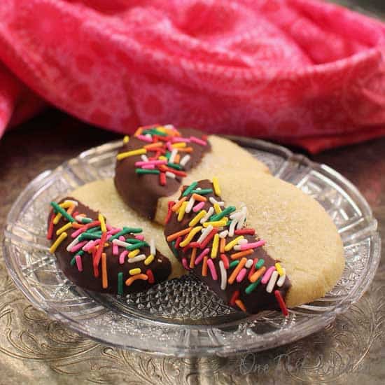 three cookies on a plate dipped in chocolate and covered with colored sprinkles next to a pink napkin.