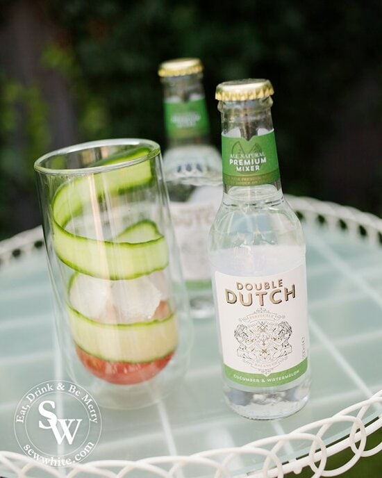 A double walled glass with a thin slice of cucumber wrapped around the glass in a swirl with fresh watermelon juice in the bottom. With a bottle of Watermelon and Cucumber Rum Punch by Double Dutch.