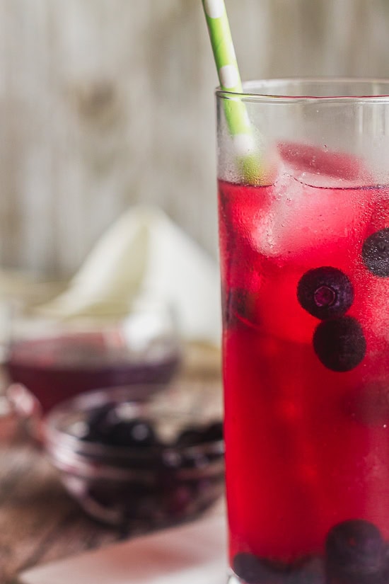 a close up of a glass of moscow mule flavored with blueberry simple syrup. There are frozen blueberries in the glass.