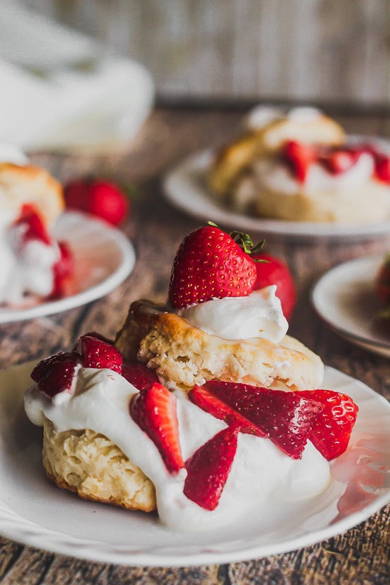 a straight on shot of a strawberry shortcake in front with other shortcakes in back, with softly whipped cream and buttermilk biscuits