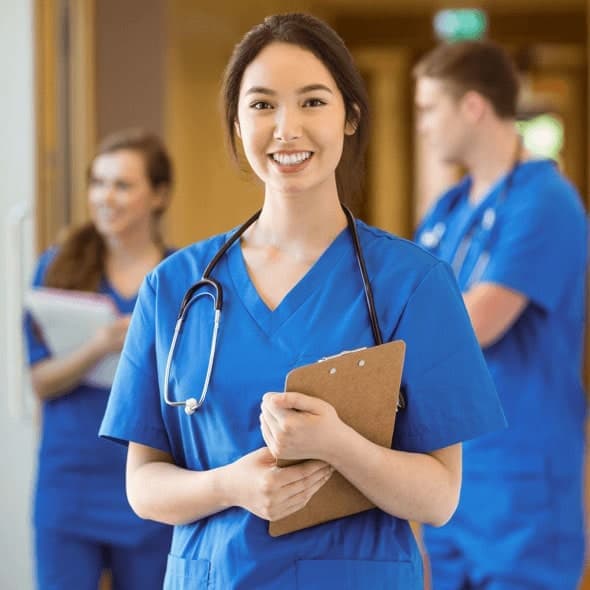 A smiling young woman in blue scrubs holding a clipboard with a stethoscope around her neck.