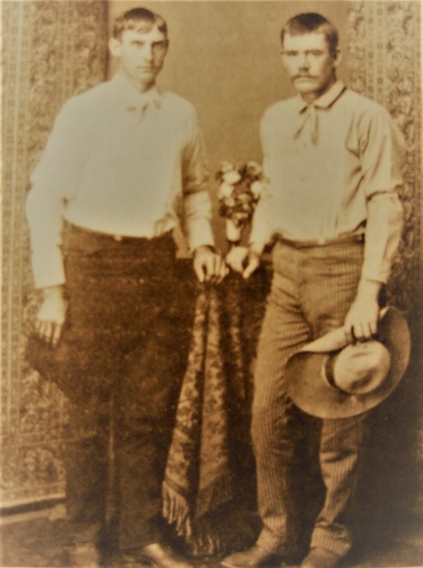Studio shot of two young men standing in cowboy boots with cowboy hats in hands.