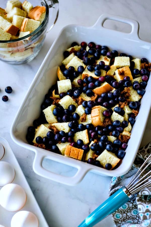 white casserole dish with bread cubes and fresh blueberries being prepped for a Blueberry Strata casserole