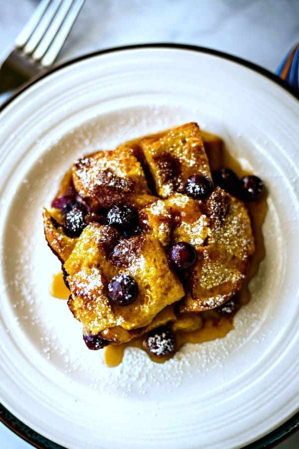 baked blueberry French toast with maple syrup on a white plate dusted with powdered sugar