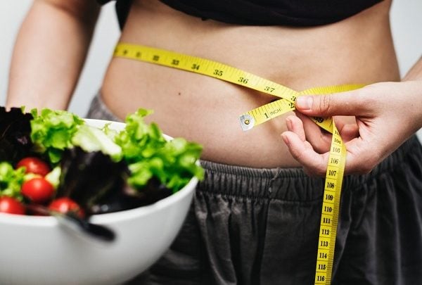 A woman holding a green salad bowl with one hand and a tape measure by her waist on one hand
