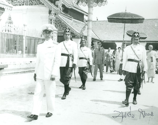 1958 black and white photo of King Rama IX in walking processional with attendants dressed in traditional Thai regalia.