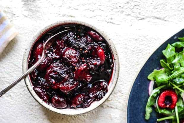 An off-white hand made owl filled with cherry ginger chutney sits on a white surface. There is a plate peaking into the corner of the image with an arugula salad and fresh cherries.