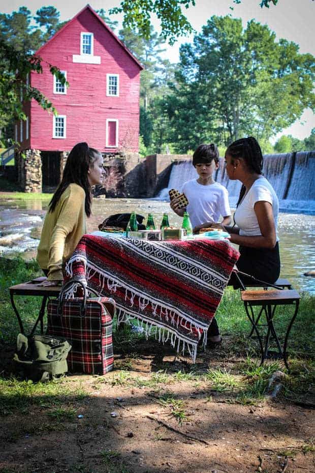 A picnic scene in fort of an old red saw mill with a water fall. There are 2 teenage girls and a small bowl at a pop up picnic table enjoying sandwiches and a game.