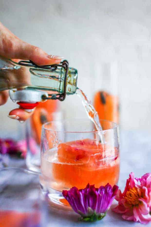 A woman is pouring water out of a glass bottle into a clear glass with a large pink hued ice cube. There are edible flowers scattered across the table top.