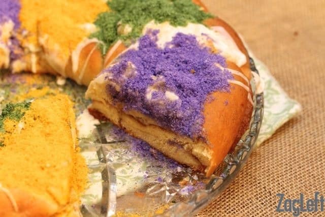 A slice taken out of a King Cake that is drizzled with white icing and topped with purple, green, and yellow sanding sugar
