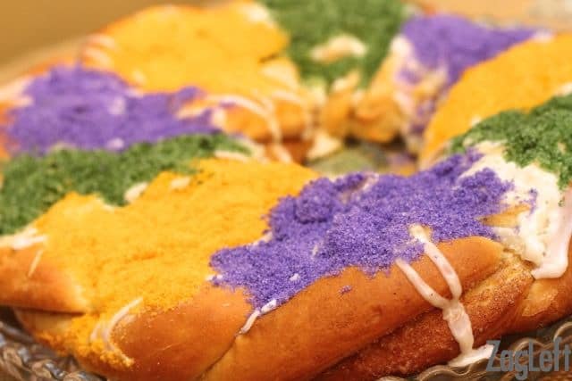 A close up of a king cake drizzled with white icing and topped with purple, green, and yellow sanding sugar