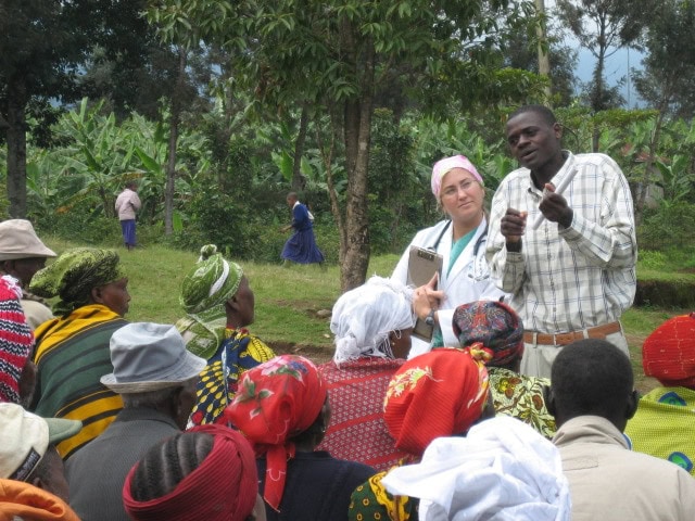 Doctor in white jacket stands next to translator and address a crowd waiting for medical services with the jungle in the background