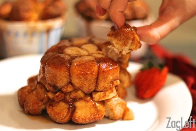 Fingers grabbing a piece of Apple Fritter Monkey Bread from a big plate garnished with strawberry slices