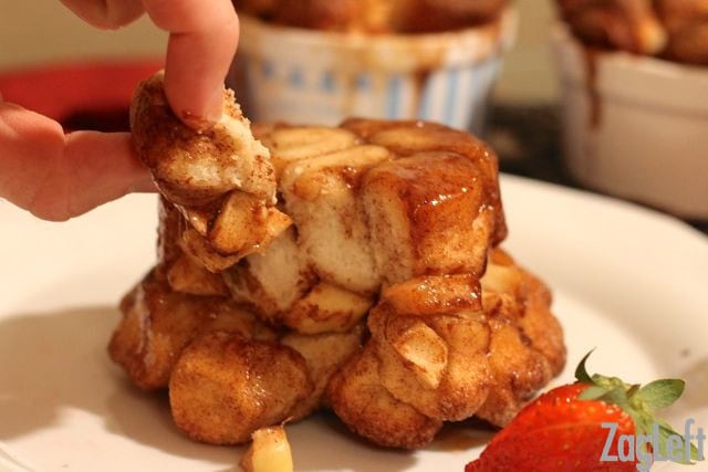Fingers grabbing a piece of Apple Fritter Monkey Bread from a big plate garnished with strawberry slices