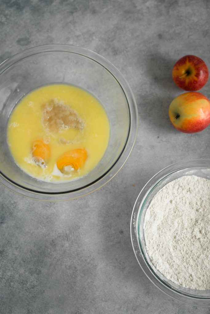 The ingredients to make apple cinnamon bread in bowls, the wet mixture ingredients in one bowl and the dried ingredients in another bowl and couple apples in the back ground