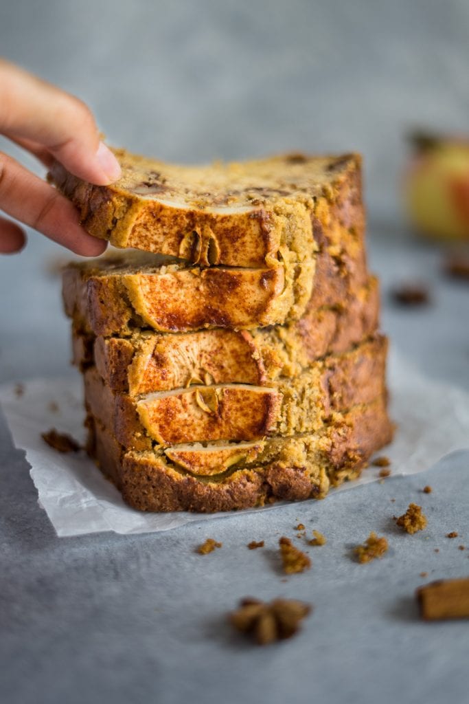 Slices of apple bread holding one with the fingers and some scrambled around and some apples in the background.