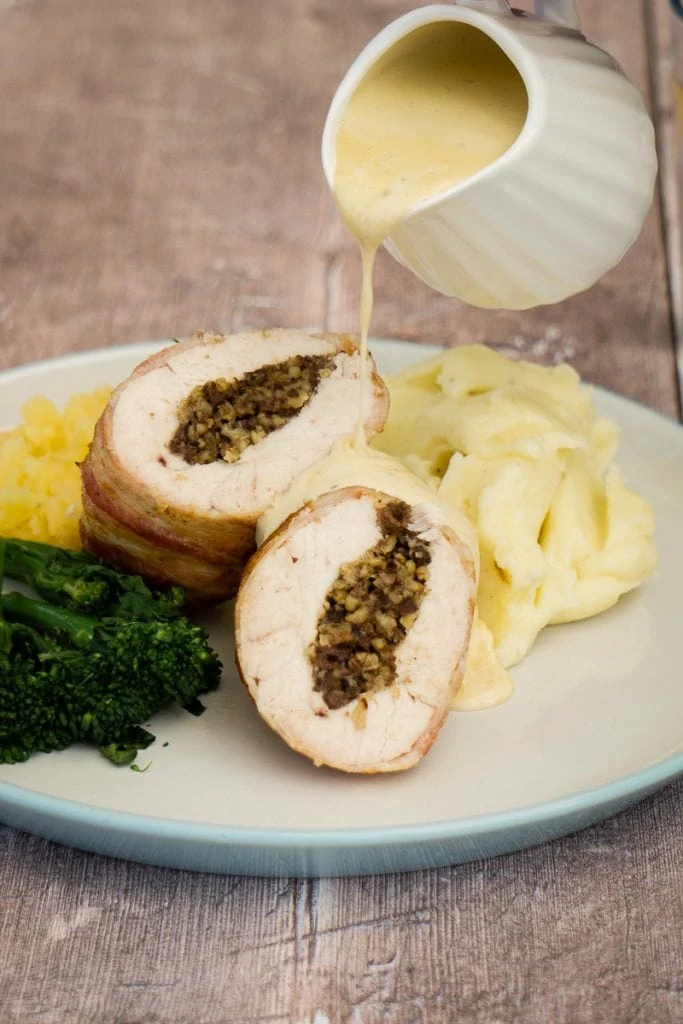 Chicken Balmoral - Chicken stuffed with haggis wrapped in bacon on a plate with vegetables and jug pouring whisky sauce over it