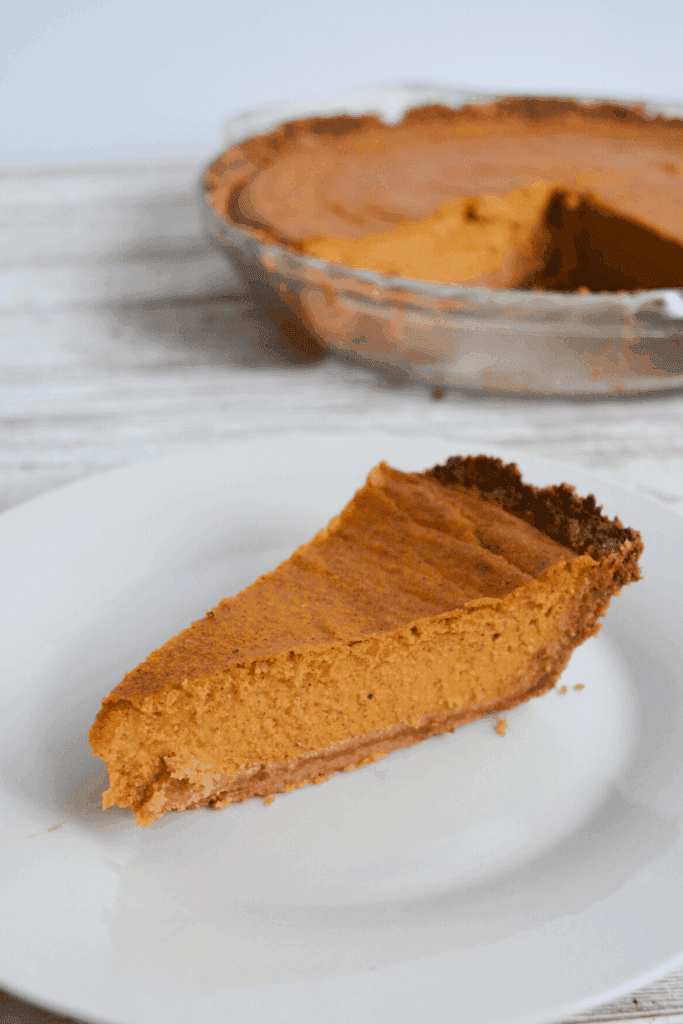 slice of pumpkin pie on white plate with full pie in the background
