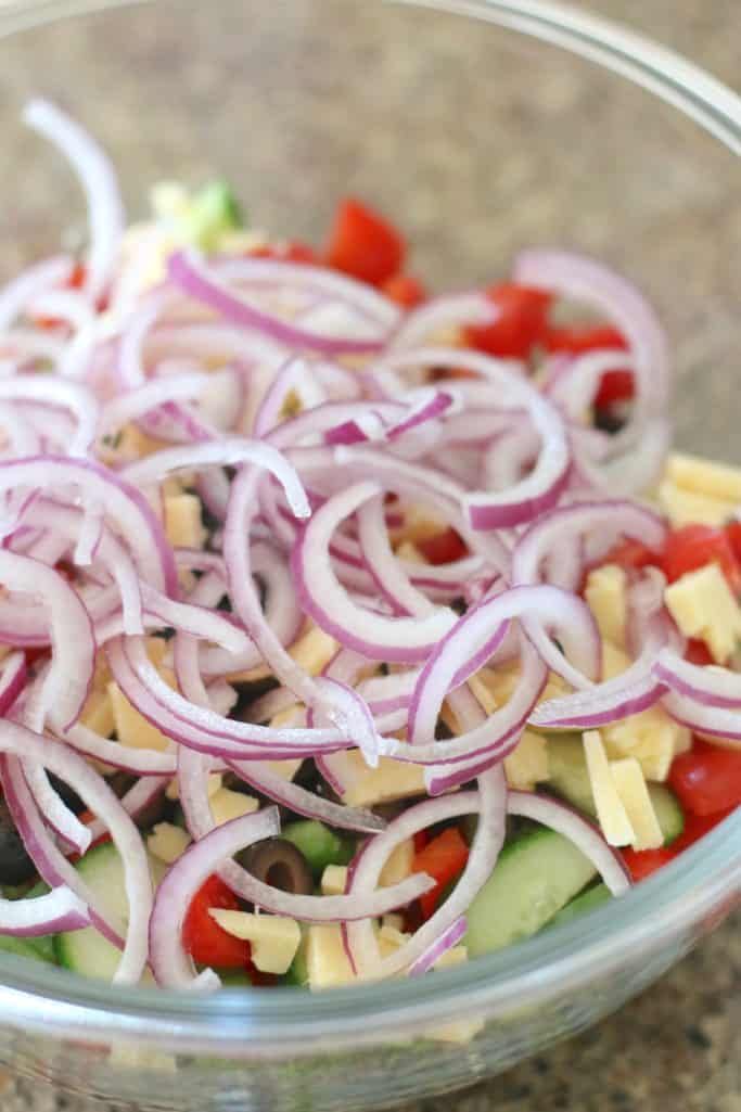 Cooked spaghetti noodles, Dubliner diced cheese, sliced red onion, sliced cherry tomatoes, green pepper, red pepper, cucumber, sliced olives in a large clear glass bowl