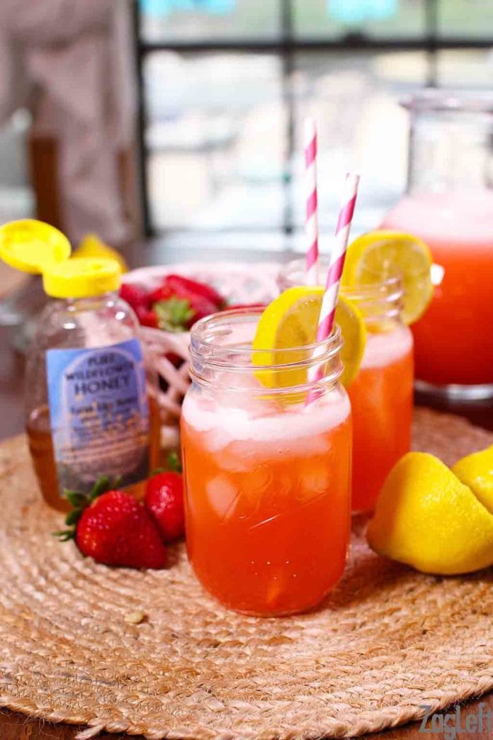 Two glasses of strawberry lemonade in mason jar glasses with a paper straw and a lemon wheel garnish next to a container of honey, two strawberries, and two lemons