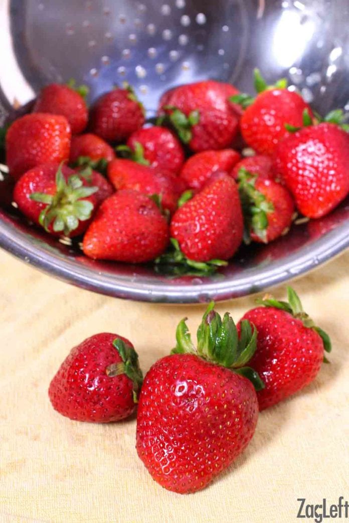 Close up of three strawberries on a cloth with a strainer of strawberries behind them