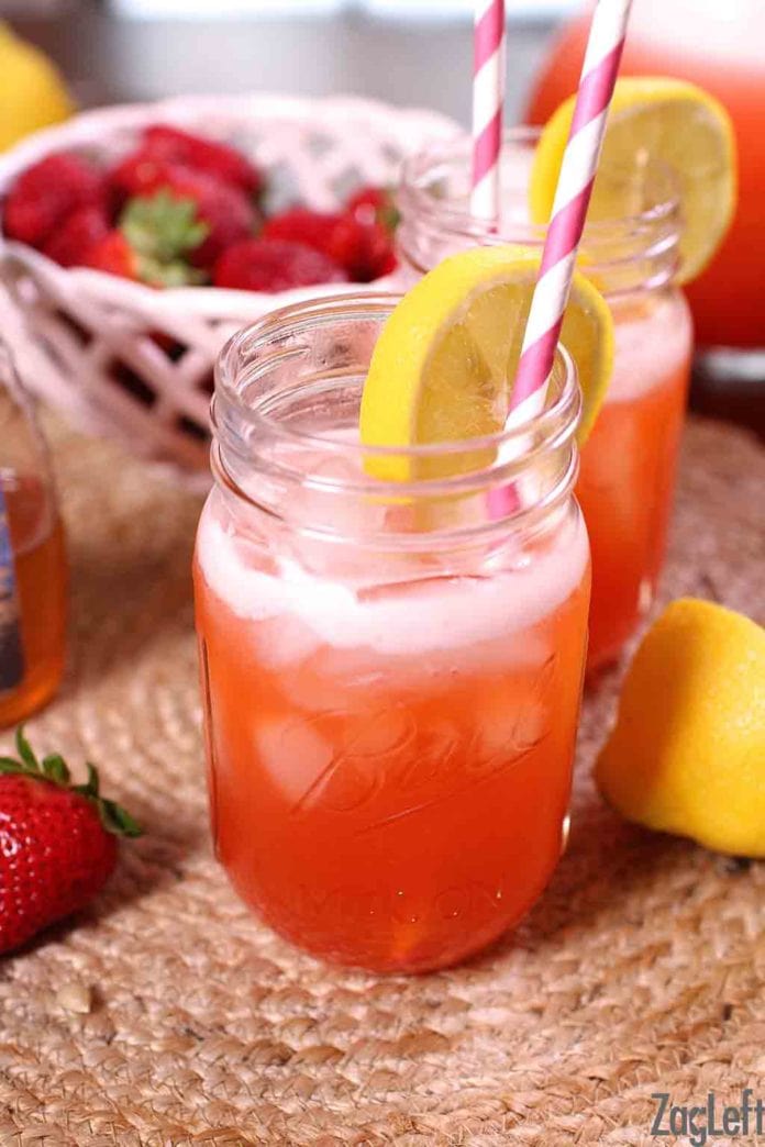 Two glasses of strawberry lemonade in mason jar glasses with a paper straw and lemon wheel garnish with a bowl of strawberries in the background