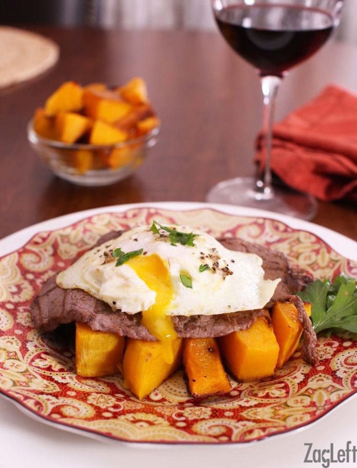 A fried egg with crispy edges and a warm yolk running down the sides of a juicy steak over a bed of seasoned roasted butternut squash on a plate with a glass of red wine and a small bowl of chopped butternut squash in the background