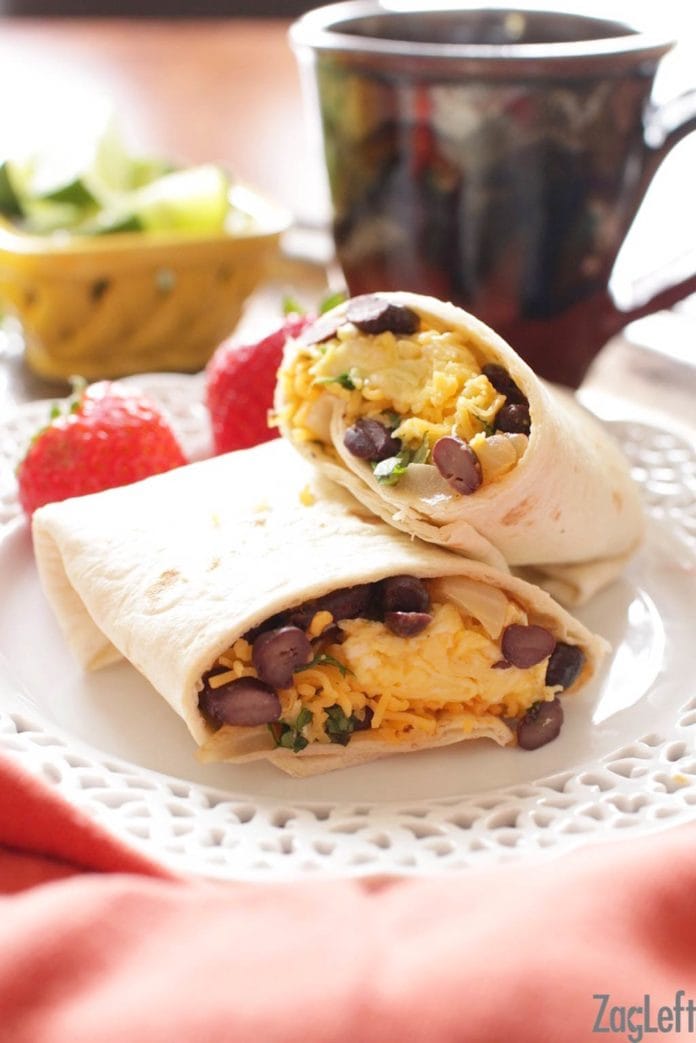 Breakfast burrito consisting of eggs, cheese, black beans, cilantro and onions cut in half on a plate with two strawberries and a coffee mug in the background