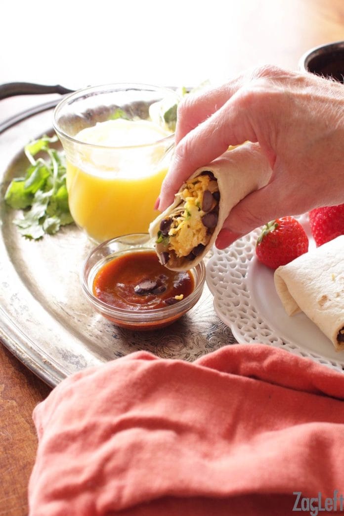 Dipping a half of a breakfast burrito into a small bowl of red salsa that's on a metal tray with a small glass of orange juice