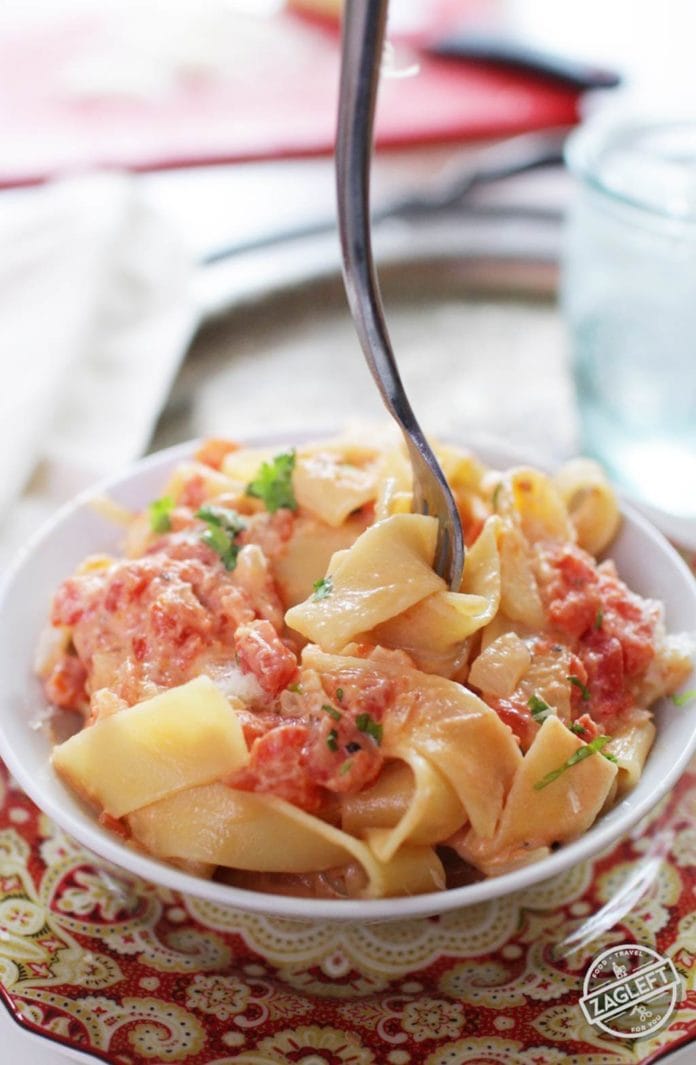 A fork in a bowl of pasta with vodka sauce plated on a metal tray with a small glass of ice water