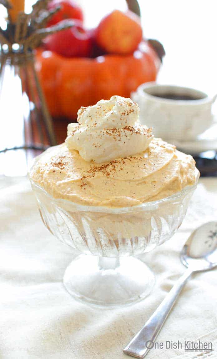 Pumpkin Cheesecake in small dessert glass topped with whipped cream with a pumpkin and a mug of coffee in the background