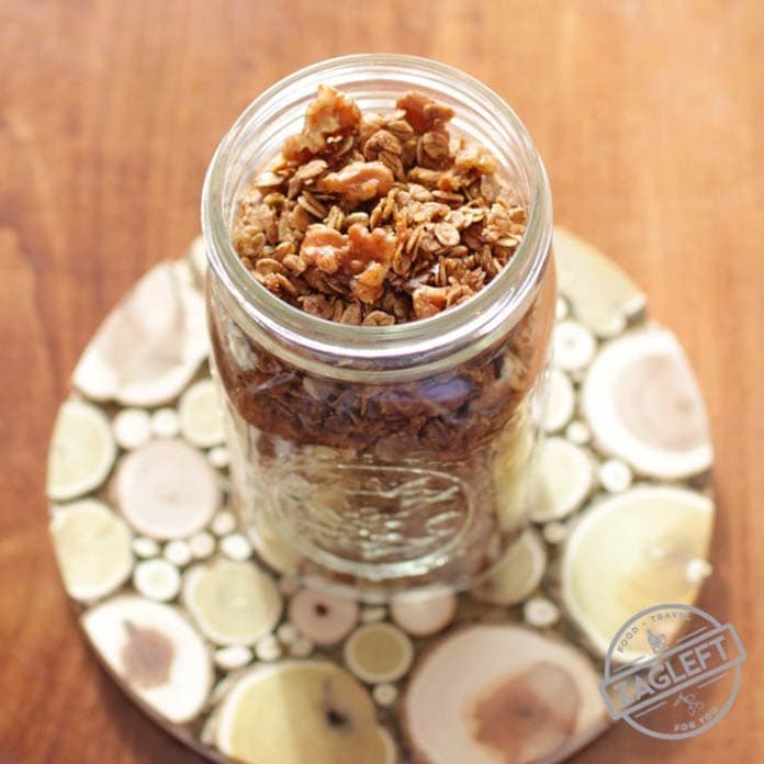 An overhead view of a jar of homemade gingerbread granola topped with walnuts on a wooden trivet